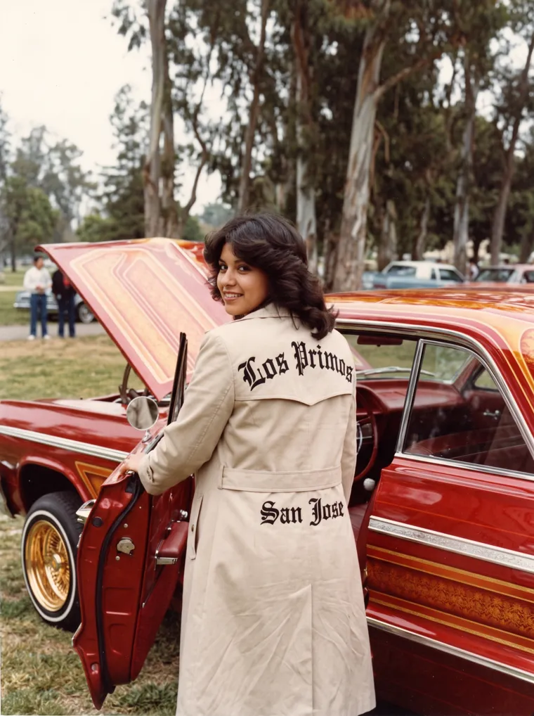 A woman in a Los Primos San Jose club jacket looks back over her shoulder at the open door of the candy-red '64, on a 1991 car magazine cover